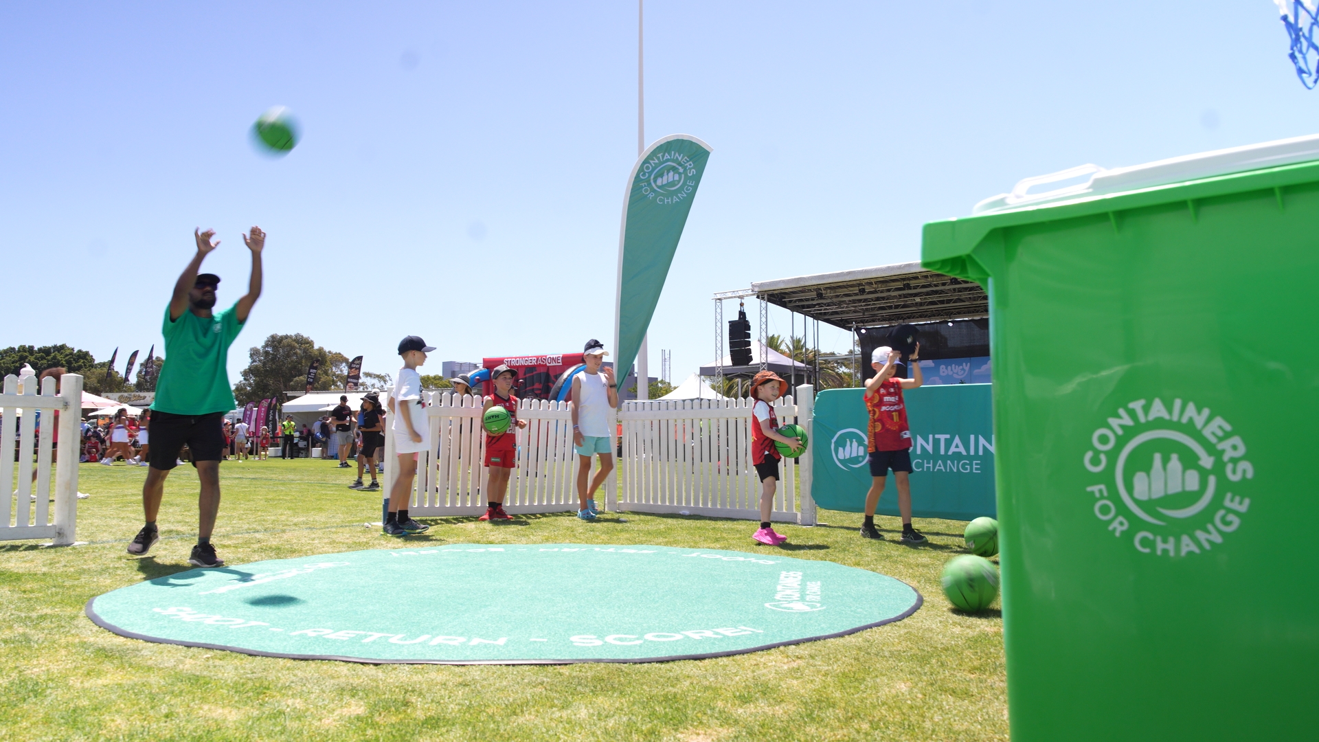 A person is throwing a green basketball in a park. A green Containers for Change wheelie bin is in the foreground.