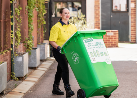 A woman with brown hair in a ponytail and wearing a yellow high-visability shirt is walking and pushing a green and white Containers for Change bin.